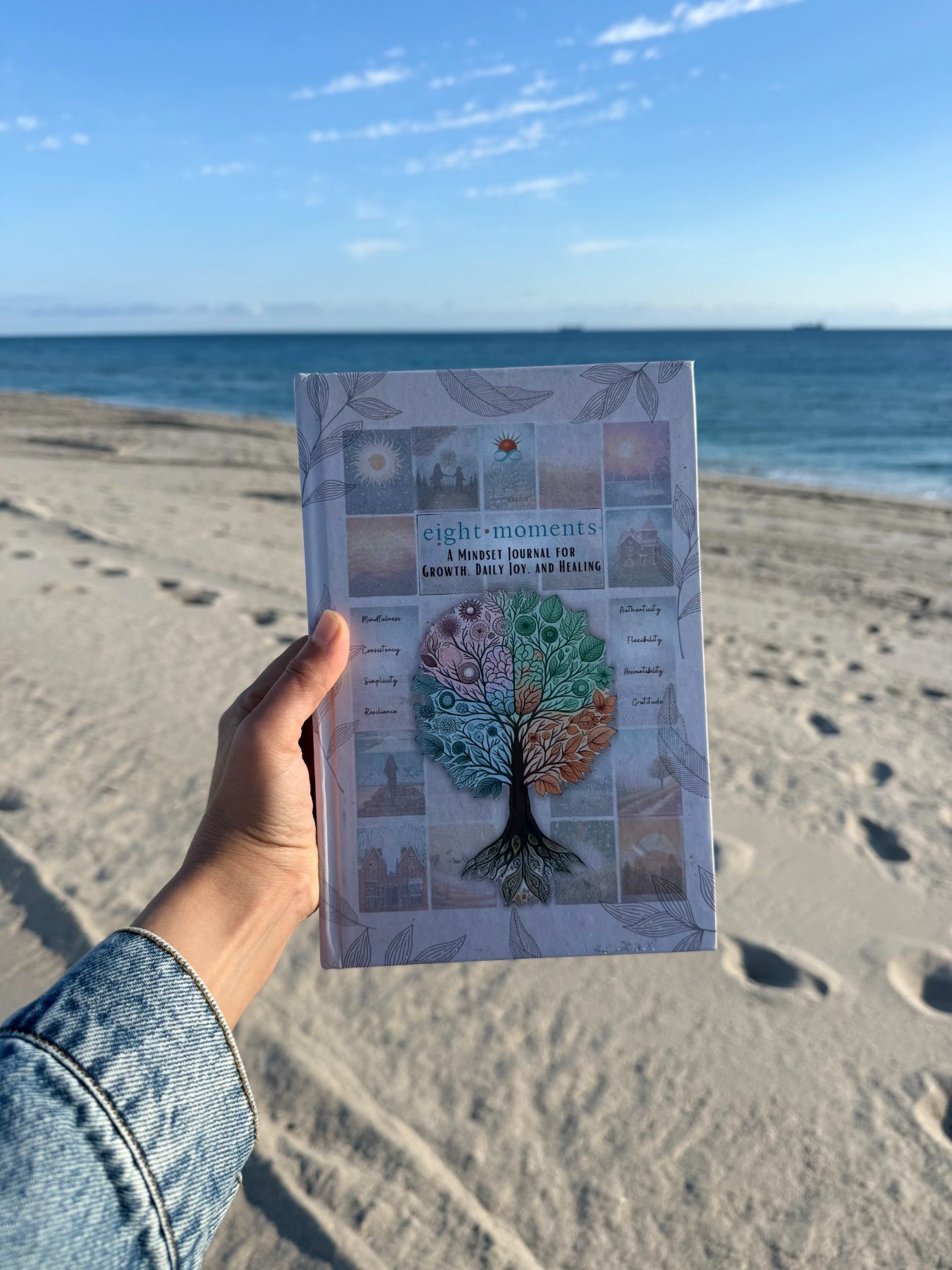 The Eight Moments journal held up against the ocean and open summer sky